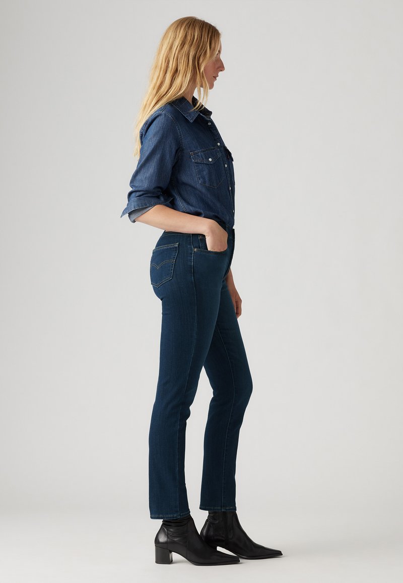Jean en denim bleu foncé au design ajusté, doté de poches, associé à une chemise en denim bleu foncé à boutons. Bottines noires à talons.