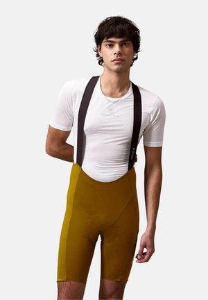 Jeune homme portant un sous-vêtement de cyclisme à manches courtes blanc et un cuissard à bretelles couleur moutarde avec des bretelles noires, debout devant un fond uni.