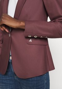 Burgundowy blazer o eleganckim designie, z srebrnymi guzikami i haftowanym logo na kieszeni. Gładka tekstura i dopasowany krój.