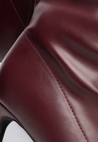 Detailní záběr hladké, tmavě vínové kožené boty s viditelným šitím a částí černého vysokého podpatku.