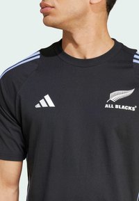 Sort kortærmet trøje med blå striber på skuldrene, hvid Adidas-logo og teksten "ALL BLACKS" i hvid med et bladdesign.