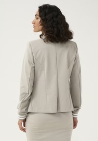 Blazer gris claro con un diseño estructurado, textura suave y puños a rayas. Presenta detalles de costura y una silueta ajustada.