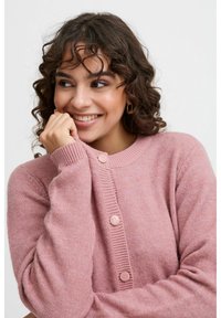 Pull en tricot rose avec un col rond, doté de poignets côtelés et de cinq boutons assortis sur le devant. Texture douce avec un léger éclat.