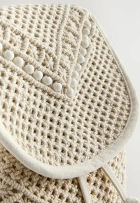 Beżowa torba z plecionego materiału o teksturowanym wzorze, z klapą ozdobioną białymi koralikami i obszytymi brzegami.