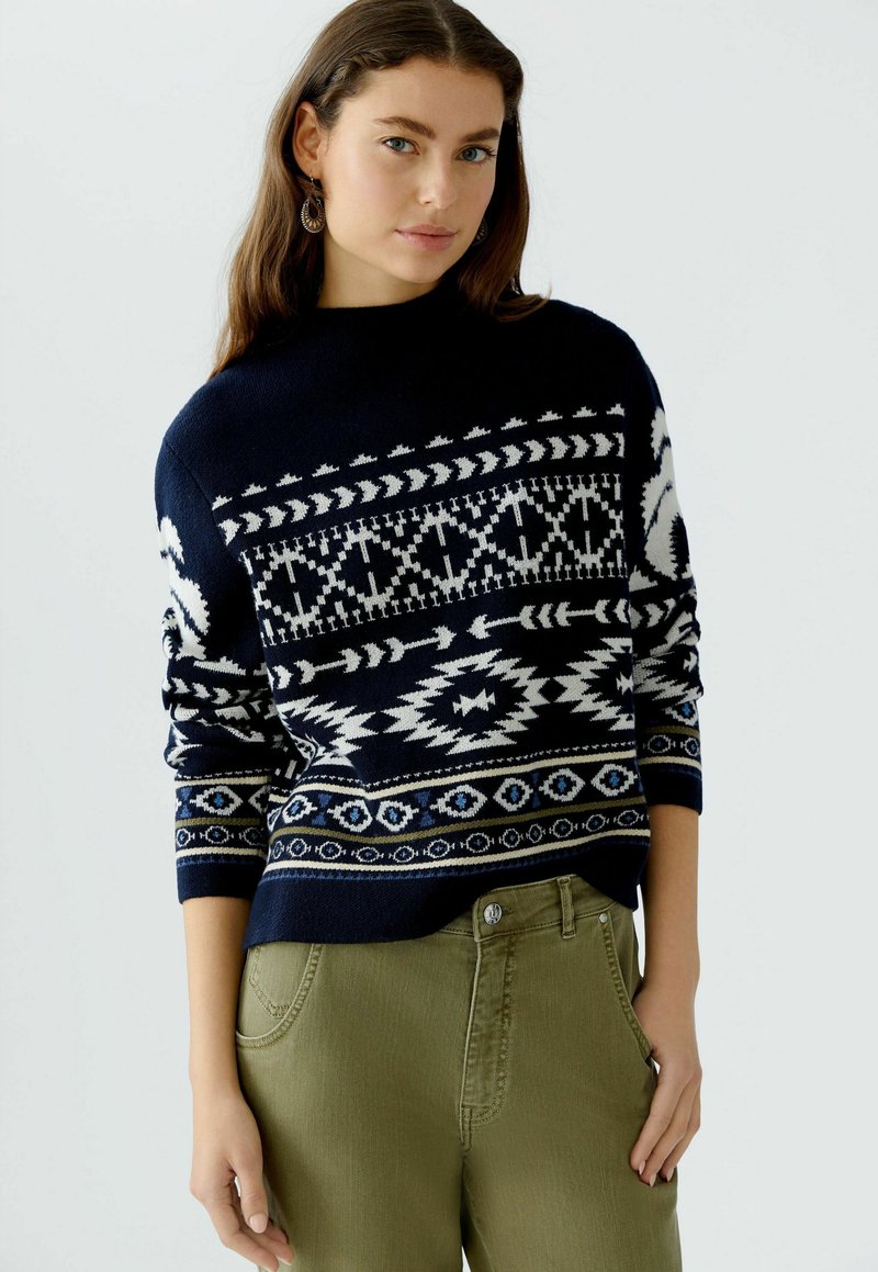 Navy trui met witte en blauwe geometrische patronen, ronde hals en lange mouwen, gecombineerd met olijfgroene broek.