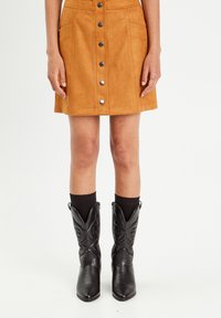 Jupe en suède beige avec une coupe évasée, fermeture à boutons sur le devant et deux poches. Associée à des bottes de cowboy noires et des chaussettes noires côtelées.