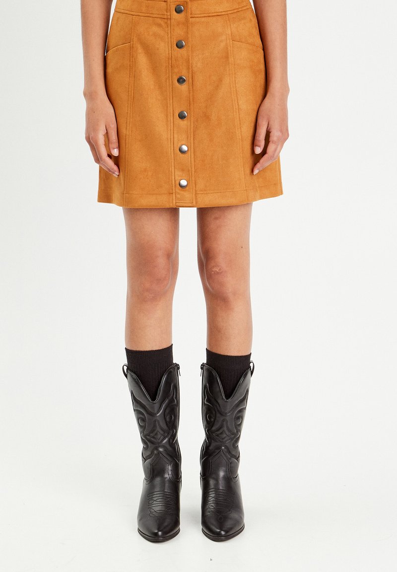 Jupe en suède beige avec une coupe évasée, fermeture à boutons sur le devant et deux poches. Associée à des bottes de cowboy noires et des chaussettes noires côtelées.