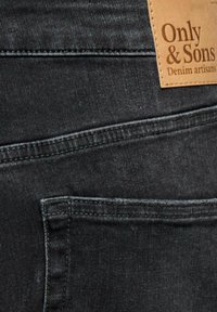 Gros plan sur la poche arrière d'un jean en denim noir avec une étiquette en cuir beige portant l'inscription Only & Sons Denim artisans cousue au-dessus.