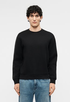 Νεαρός άνδρας με σκούρα σγουρά μαλλιά, φορώντας ένα απλό μαύρο φούτερ με στρογγυλή λαιμόκοψη και μπλε τζιν, στέκεται μπροστά σε ανοιχτό γκρι φόντο.