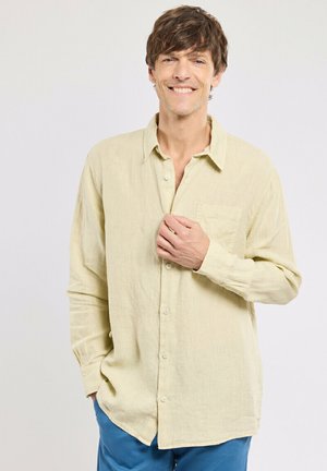 Homme souriant, portant une chemise beige à boutons et un pantalon bleu, avec une main dans la poche et l'autre ajustant le col de la chemise.