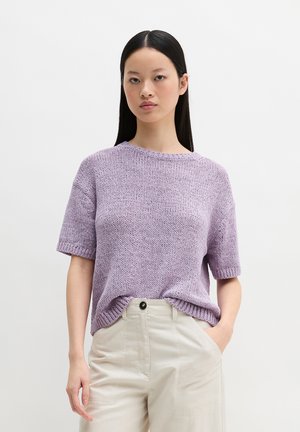 Jonge vrouw met lang zwart haar, gekleed in een lila gebreide trui met korte mouwen, ingestopt in een beige broek met hoge taille, staand tegen een effen achtergrond.