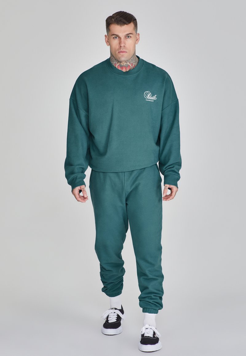 Sudadera verde y pantalones de chándal a juego hechos de una tela suave. Presenta un logo en el pecho, puños acanalados y cinturilla elástica.