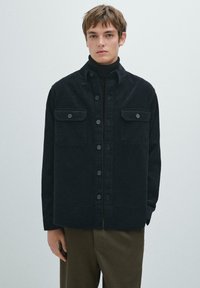 Zwarte corduroy overhemdjas met twee borstzakken, knoopsluiting en een gestructureerde afwerking. Draagt over een zwarte coltrui.