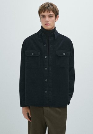 Sort corduroy skjortejakke med to brystlommer, knaplukning foran og en struktureret overflade. Bæres over en sort poloshirt.