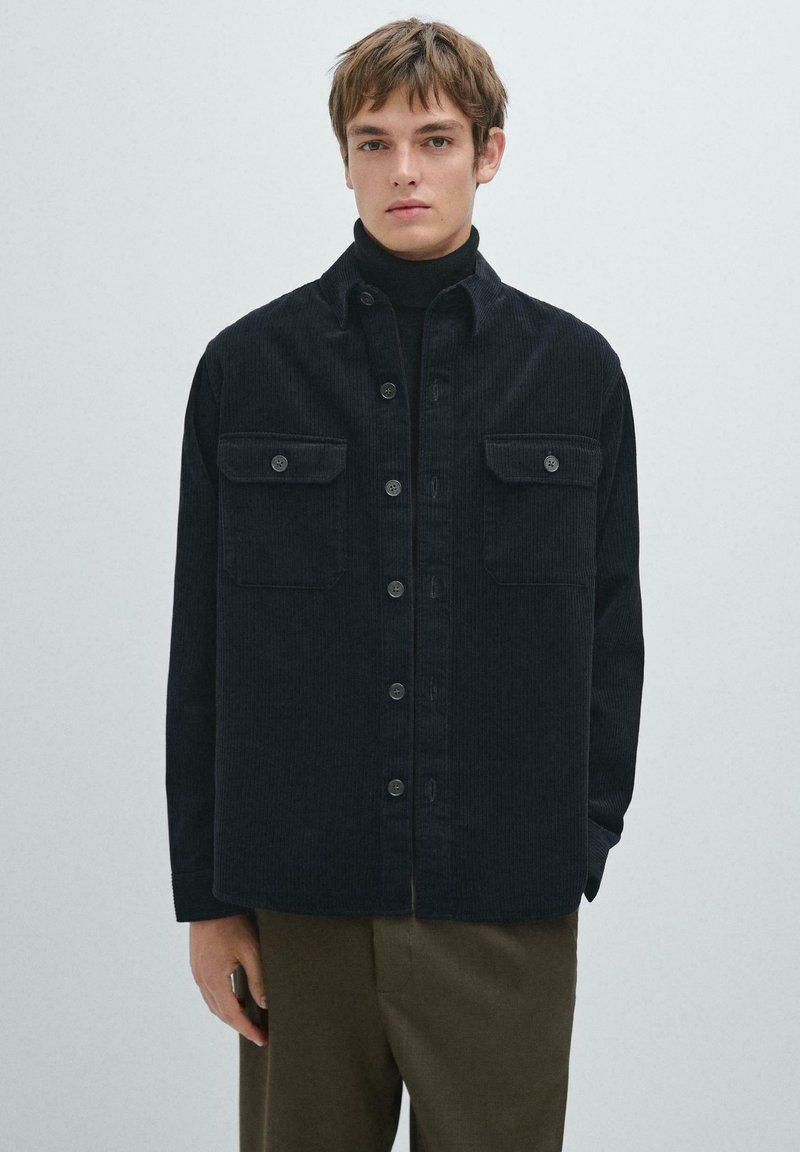 Zwarte corduroy overhemdjas met twee borstzakken, knoopsluiting en een gestructureerde afwerking. Draagt over een zwarte coltrui.