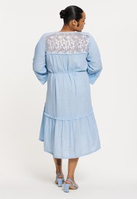 Hellblaues Chambray-Kleid mit gestuftem Rüschensaum, langen Ärmeln und Spitzenrückenverzierung. Abgerundet mit hellblauen Blockabsätzen.