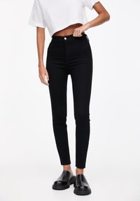 Jeans skinny de talle alto en negro con una textura suave, diseño de cinco bolsillos y cierre de botón de metal, combinados con una camisa corta blanca.