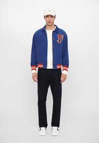 Blue corduroy jacket with red and white striped cuffs, featuring an embroidered logo patch, worn over a white sweater with dark jeans.