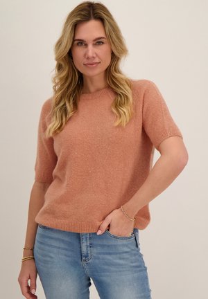 Vrouw met golvend blond haar, gekleed in een perzikkleurige trui met korte mouwen en lichtblauwe jeans, poseert met één hand in de zak tegen een effen achtergrond.