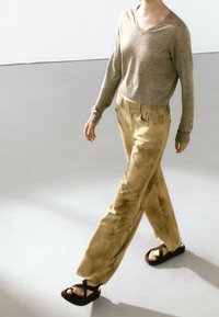 Pull en tricot beige à col en V, associé à un pantalon droit légèrement texturé couleur marron clair et des sandales plates noires.