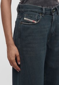 Gros plan d'une personne portant un jean Diesel bleu foncé avec une petite étiquette visible sur la poche avant droite et la main gauche reposant à proximité.