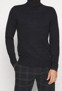 Man som bär en svart polotröja med ribbade mudd och nederkant, i kombination med mörka rutiga byxor, med händerna i fickorna.