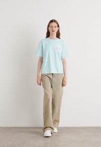 Lichtblauw katoenen t-shirt met korte mouwen, met een logo op de linkerborst. Gecombineerd met beige wijde broek en witte sneakers.