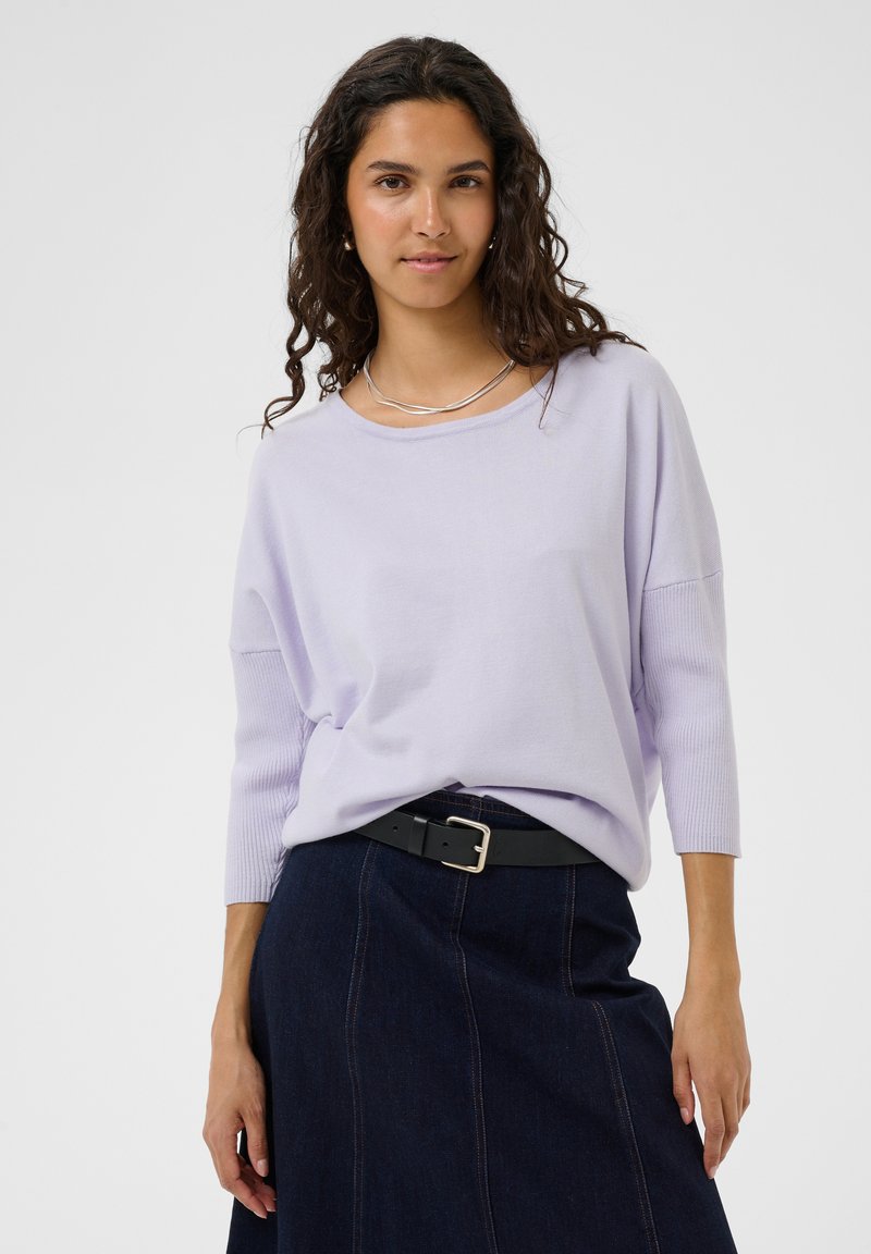 Femme portant un haut à manches longues violet clair, une jupe en jean foncé, une ceinture noire et un collier en argent, debout devant un fond gris clair.