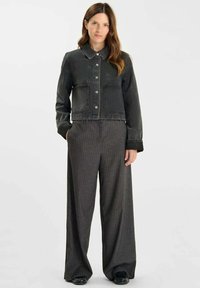 Mujer vestida con una chaqueta corta de botón negra y pantalones de pinzas de talle alto y pierna ancha, con las manos en los bolsillos, de pie frente a un fondo sencillo.