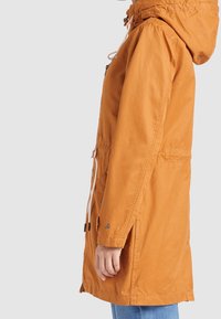 Pomarańczowa lekka kurtka z kapturem na sznurek i talią. Posiada boczne kieszenie oraz guzik jako detal na rękawie. Gładka tkanina.
