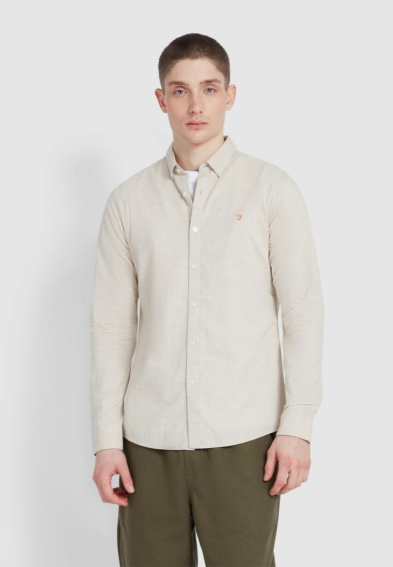 Chemise boutonnée beige clair en lin avec une texture douce, ornée d'un petit logo brodé orange et de boutons blancs.