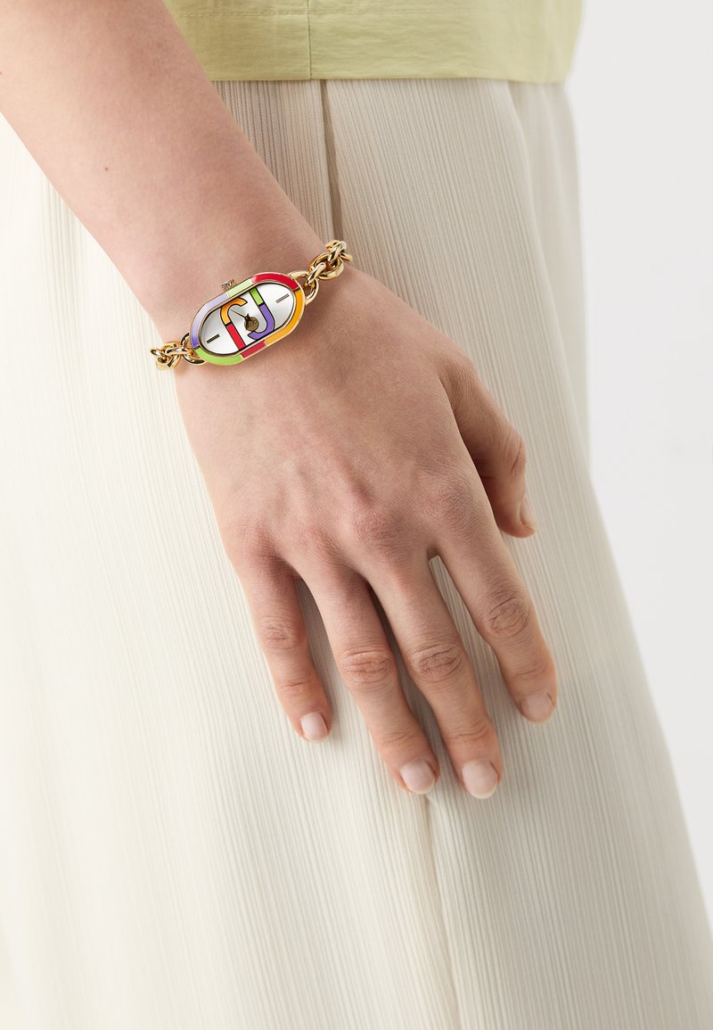 Bracelet en or avec un charm ovale abstrait coloré comportant des sections rouges, roses, vertes et bleues. Porté sur un poignet fin, avec un fond texturé.