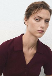 Polo shirt bordeaux à manches courtes avec col, doté d'une fermeture à boutons et d'une texture tricot lisse, sur fond à texture claire.