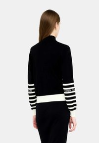 Cardigan nero lavorato a maglia con un alto collo, con strisce orizzontali bianche sulle maniche e sull'orlo. Accenti del logo bianco sui dettagli a strisce.