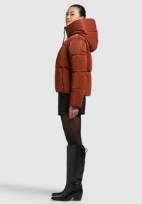 Oranžinė paminkštinta striukė su aukštu apykakliu ir gaubtu, turinti priekinį užtrauktuką ir šonines kišenes, dėvima su juoda suknele ir aukštomis juodomis ilgaaulėmis.