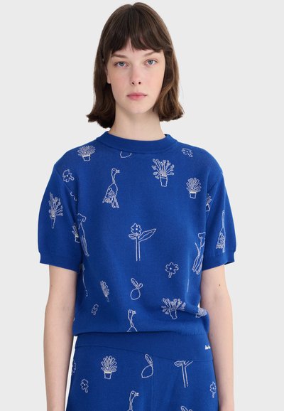 Kobieta ubrana w niebieski sweter z krótkim rękawem i pasujące spodnie ozdobione białymi rysunkami roślin i ptaków.