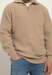 Maglione beige lavorato a maglia con colletto a mezza zip, texture a coste e maniche lunghe, abbinato a jeans blu e una tasca frontale visibile.