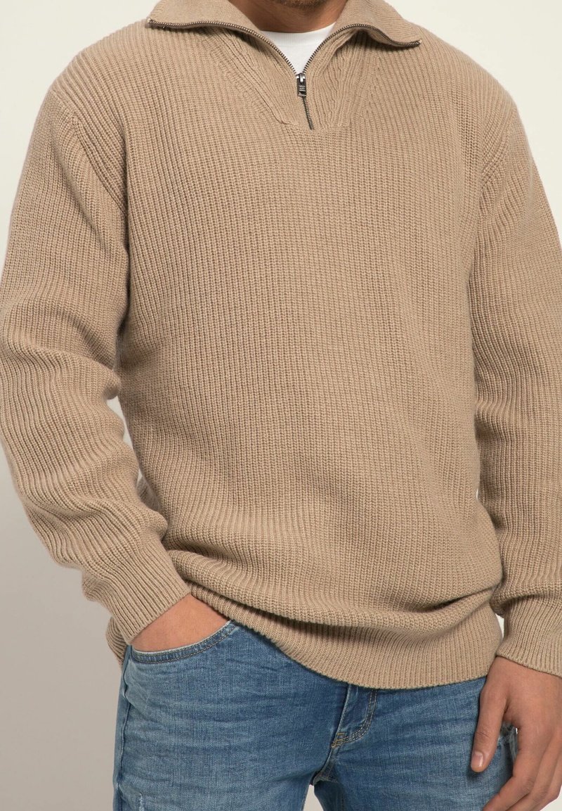 Maglione beige lavorato a maglia con colletto a mezza zip, texture a coste e maniche lunghe, abbinato a jeans blu e una tasca frontale visibile.