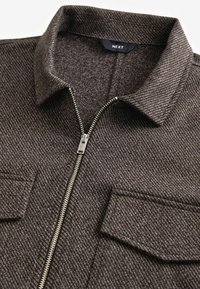 Bruine textuurige jas met ritssluiting, een kraag, twee zakken aan de voorkant en een metalen rits. De stof heeft een subtiel diagonaal patroon.