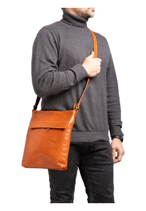 Man draagt een grijze coltrui en zwarte spijkerbroek, en heeft een bruine leren crossbodytas over zijn schouder hangen.