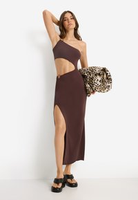 Καφέ φόρεμα με ένα ώμο, με ψηλή σχισμή και μεταλλικές λεπτομέρειες. Το μοντέλο κρατάει ένα παρεό με λεοπάρ σχέδιο και φοράει μαύρα σανδάλια με φαρδύ τακούνι.