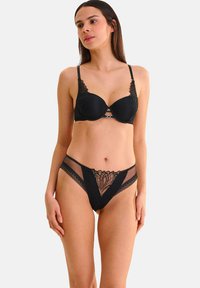 Ensemble de lingerie noire comprenant un soutien-gorge rembourré avec des accents en dentelle et des panneaux transparents, associé à une culotte bikini assortie avec des motifs décoratifs.
