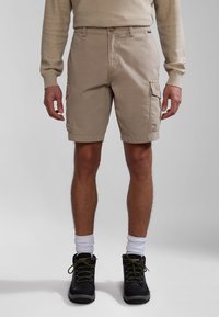 Pantalones cortos de cargo beige con bolsillos laterales, tejido duradero y cierre de botón. Llevado con zapatos negros y calcetines blancos.