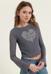 Grå, kortklipt langermet strikket topp med åpen vevtekstur og hjerte mønster foran. Utsvingte ermer forsterker design detaljene.