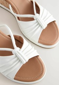 White leather sandals feature a knotted design at the vamp, a cushioned brown insole, and a subtle wedge heel with a textured cork accent.