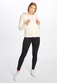 Mujer de pie con las manos cerca del cuello, vistiendo una sudadera con cremallera blanca y esponjosa, pantalones ajustados negros y zapatillas blancas sobre un fondo blanco liso.