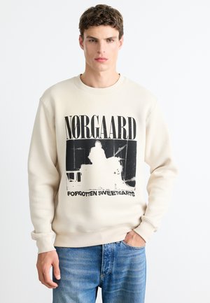 Man wearing beige sweatshirt with black graphic and text "Norgaard Forgotten Sweethearts," paired with blue jeans, standing against white background.