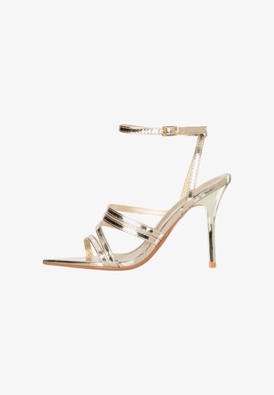 Sandale à talons hauts dorée avec fines lanières, une fine bride chevillère avec boucle, bout ouvert et talon aiguille fin sur fond blanc.