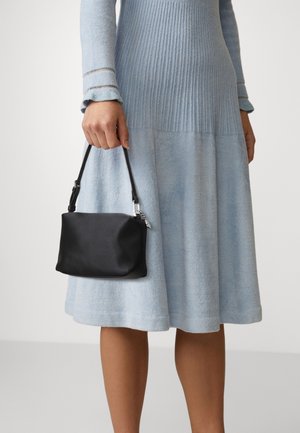 Femme portant une robe en maille à manches longues bleu clair, tenant un petit sac à main noir avec une courte bandoulière à côté d'elle.
