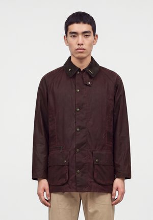 Jeune homme portant une veste marron foncé boutonnée avec deux poches avant et un pantalon beige sur un fond clair uni.